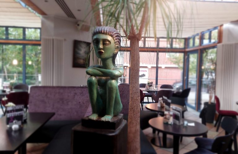 Blick in den Gastraum im Wintergarten im Café Sculptura in Altenberge Grüne Statue einer sitzenden Figur in einem Café mit Palmen und modernen Möbeln.